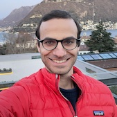 Matthew Mario Di Pasquale is smiling at the camera. His head is facing the camera, but his chest is angled slightly to his left. His hair is short and combed to his right. He has a day's worth of stubble. He's wearing round glasses with black frames and a red Patagonia jacket over a navy blue sweater. Behind him is a view of rooftops, trees, a lake, houses, mountains, and a hazy light blue sky.