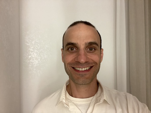 Matthew Mario Di Pasquale is facing and smiling at the camera showing his upper teeth. His hair is short. He has a couple days of stubble. He's wearing an ecru collared shirt and an ecru t-shirt underneath. Right behind him is an off-white wall and the edge of a beige curtain.