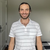 Matthew Mario Di Pasquale is smiling at the camera. His head and torso are facing the camera, and his arms are at his sides. His hair is short with curls around his ears and combed to his right. He has a two-week beard. His upper chest hair is visible. He's wearing a white polo shirt with thin black horizontal lines. Behind him to his right is a closed white door with a sliver handle and deadbolt. Directly behind him is an off-white wall and a brown floor. To his left is a bed covered with a black and white buffalo checked quilt.