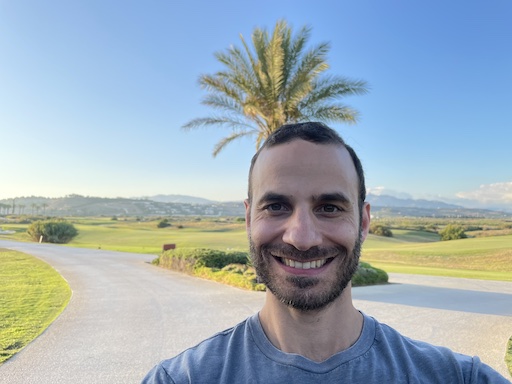 Matthew Mario Di Pasquale is facing and smiling at the camera. His hair is short. He has a three-week beard. He has on a gray shirt. Behind him is a paved road, a palm tree, a golf course, villas, mountains, and a clear blue sky.