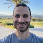 Matthew Mario Di Pasquale is facing and smiling at the camera. His hair is short. He has a three-week beard. He has on a gray shirt. Behind him is a paved road, a palm tree, a golf course, villas, mountains, and a clear blue sky.